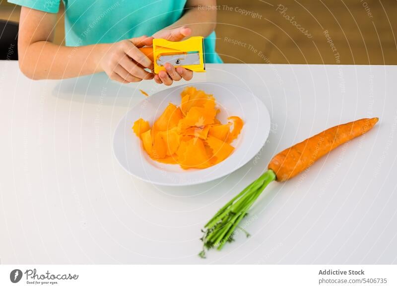 Anonymous boy standing at table with fresh carrot against blurred kitchen kid slice plate help sharpener busy child home childhood vitamin nutrition casual