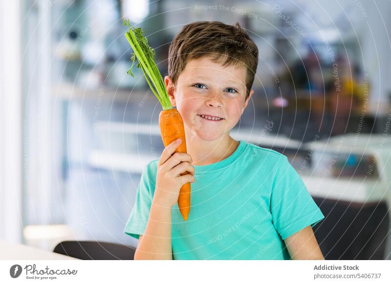 Smiling boy with carrot against blurred kitchen smile ripe healthy food child cute adorable positive yummy nutrition tasty sweet organic appetizing meal raw