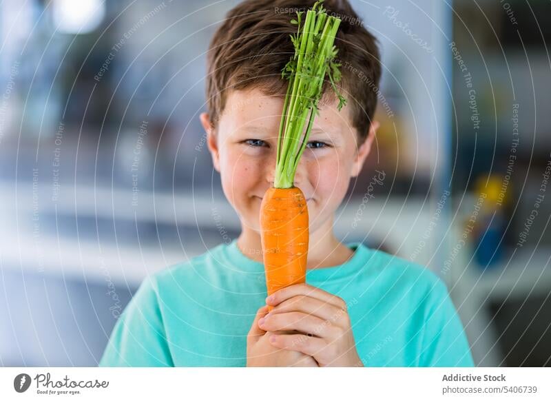 Smiling boy showing carrot against blurred kitchen smile ripe healthy food child cute adorable positive yummy nutrition tasty sweet organic appetizing meal raw