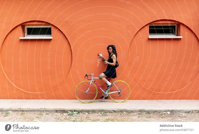 Young ethnic woman sitting on bicycle and browsing smartphone near orange wall street using sidewalk online internet summer young female mobile gadget device
