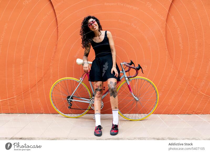 Young ethnic woman leaning against a bicycle sidewalk street orange wall building transport summer female young hispanic smile pavement stroll sunglasses
