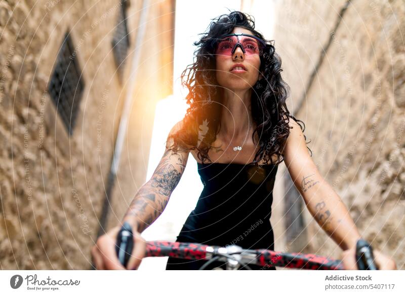 Stylish tattooed young ethnic woman standing in narrow street with bicycle confident brick wall building appearance transport summer female hispanic sunglasses
