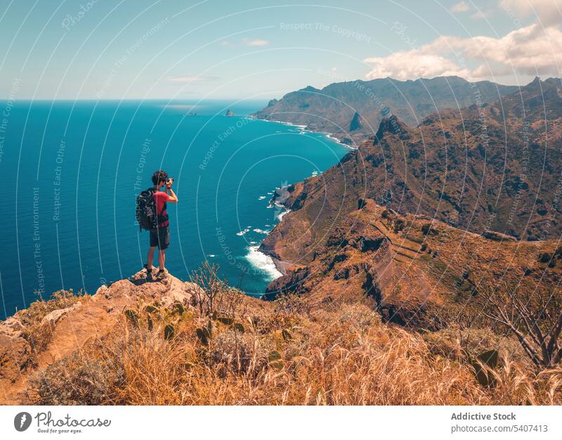 Young man with backpack standing on cliff and taking pictures with camera mountain take photo traveler hiker nature sea landscape adventure male trip tourism