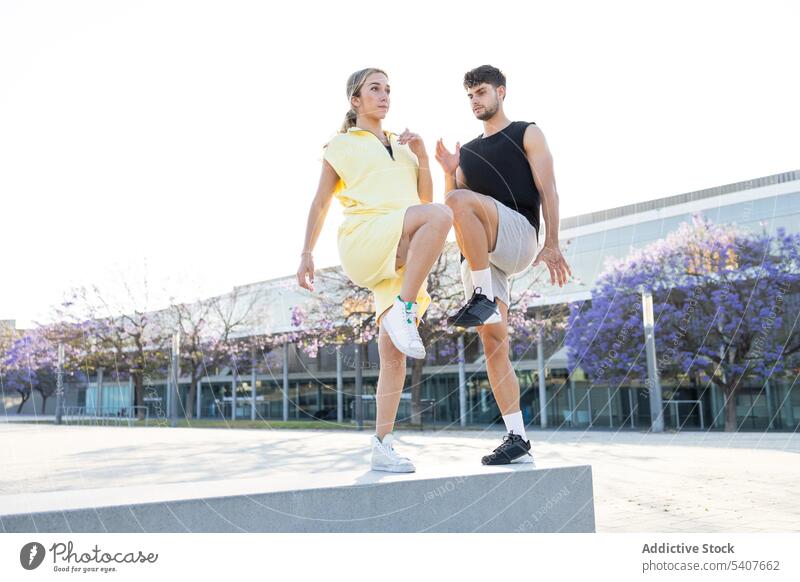 Stylish young couple doing leg up exercise on street jump training warm up workout sports ground active serious sporty sportswear sportsman together fitness