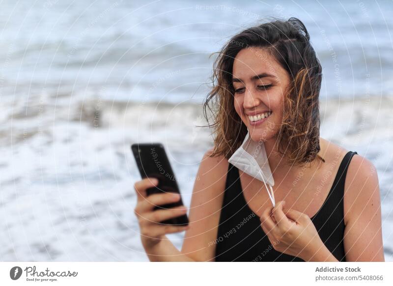 Cheerful female traveler chatting smartphone near wavy sea ocean internet online cheerful mask covid using gadget woman device surfing cellphone browsing