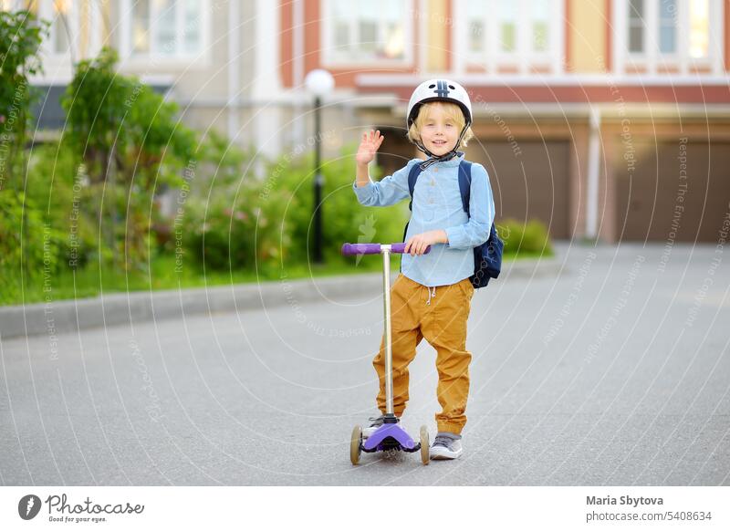 Little child in safety helmet riding scooter to school. Preschooler boy waving hand saying hi. Safety kids by way to school. Back to school concept. schoolboy
