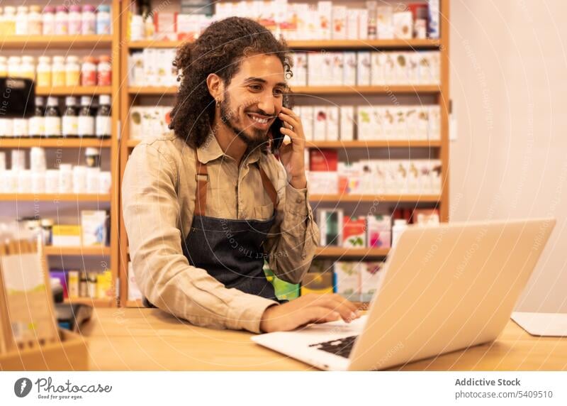 Smiling young man talking on smartphone and browsing laptop in shop smile positive communicate gadget drugstore conversation happy job speak mobile computer