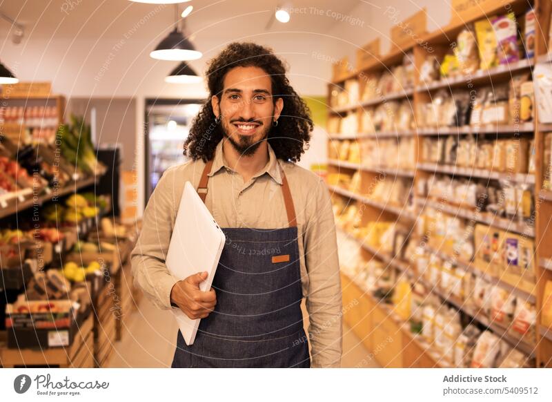 Cheerful ethnic man with laptop in grocery store smile shop positive work apron job gadget happy casual beard small business modern glad cheerful using computer