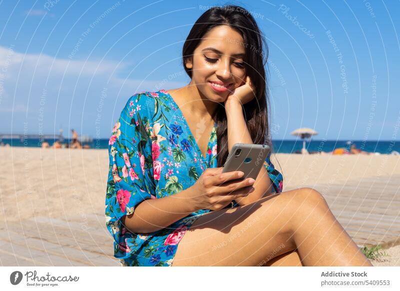 Smiling young ethnic woman sitting on beach and browsing smartphone against blue sky smile portrait using happy positive long hair female gadget device mobile