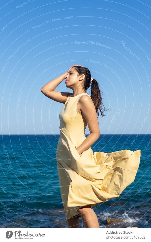 Indian female in dress enjoying summer vacation near sea woman beach shore tourist touch hair blue sky leisure carefree freedom horizon touch head