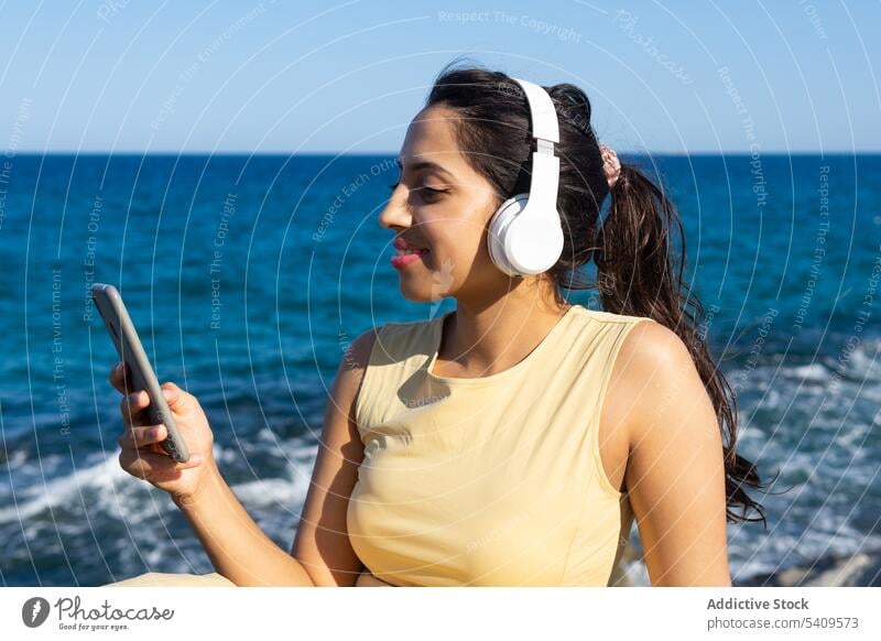 Indian woman in headphones using smartphone near sea listen browsing music smile beach enjoy seashore rest cheerful gadget device mobile surfing online relax