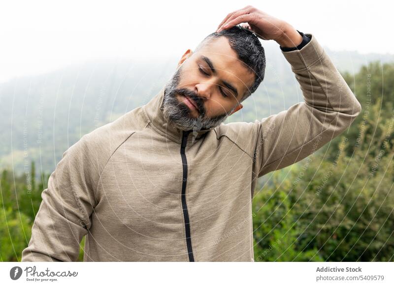 Young ethnic man standing and touching tilted head with hand during warm up eyes closed touch head meadow exercise summer nature countryside male young beard