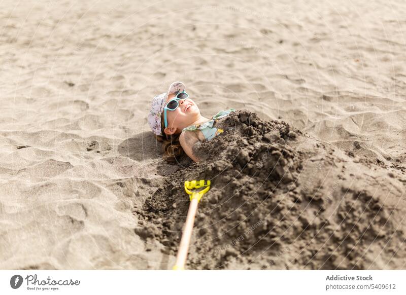 Happy kid in cap and sunglasses lying on ground with wet sand on body child beach carefree having fun playful smile summer girl recreation sunlight holiday