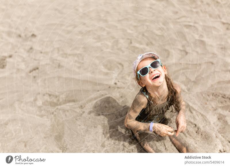 Cheerful kid in sunglasses sitting on sandy beach in cap in daylight child enjoy vacation smile swimwear summer girl seashore sunlight holiday happy chill