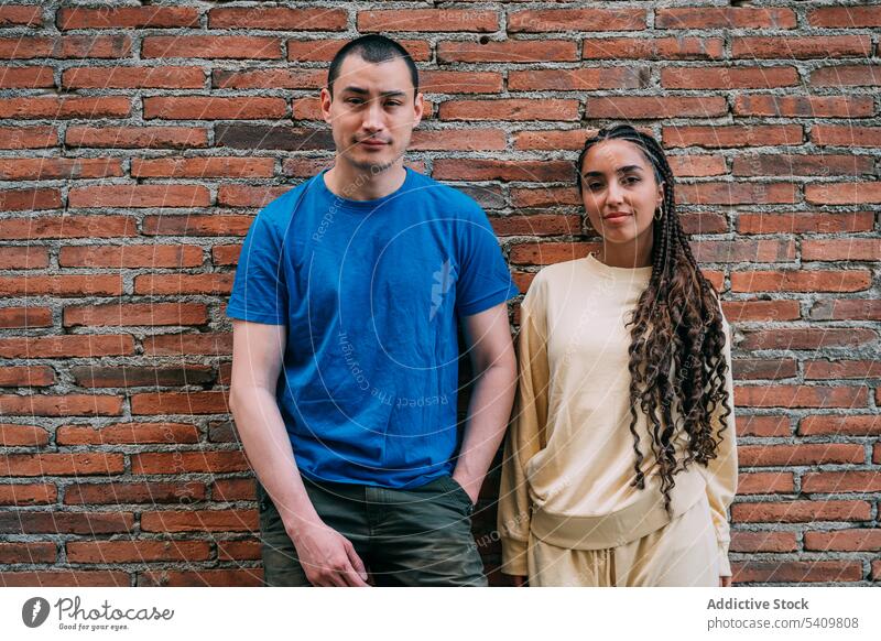 Positive couple standing near wall man woman smile brick wall street style appearance outfit together happy braid positive hispanic joy diverse multiracial