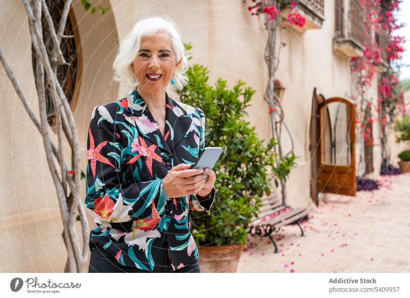Smiling senior woman standing on sidewalk and using smartphone building smile doorway online plant exterior female browsing surfing device gadget house positive
