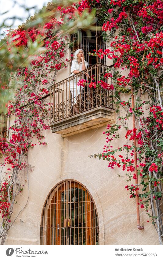Senior woman standing in balcony with iron fence near green leaves tree with flowers bloom building alone house exterior calm female senior aged leaf flora