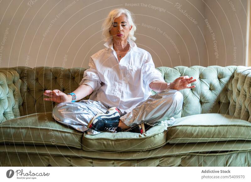 Senior woman sitting on sofa with crossed legs and doing Padmasana pose yoga meditate lotus mudra legs crossed practice eyes closed mindfulness at home female