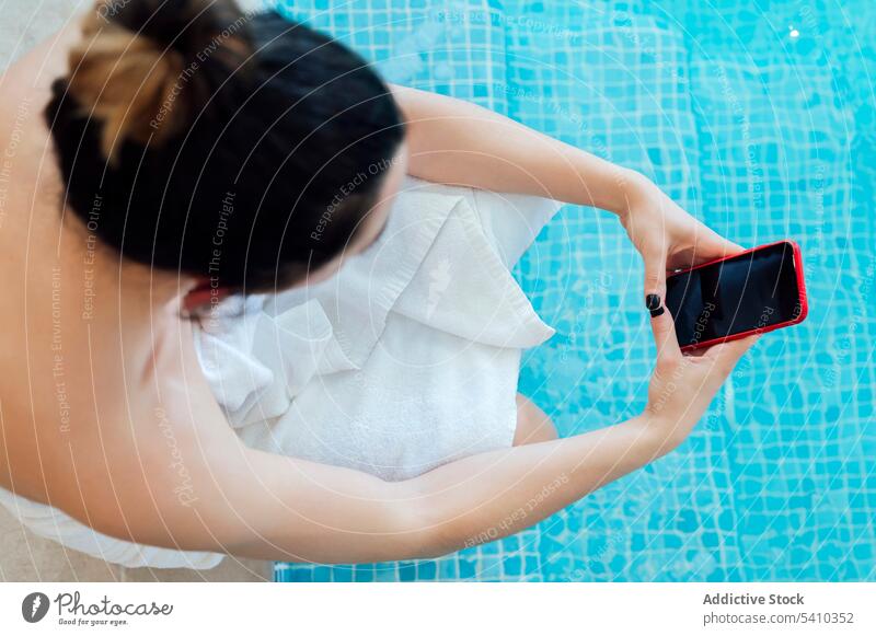 Woman in white towel sitting by pool while browsing smartphone woman vacation using poolside rest device bathrobe resort hotel relax gadget female holiday