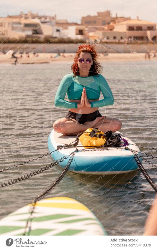 Happy woman sitting in easy yoga pose on paddleboard in sea float easy pose rest serious recreation water summer resort ocean relax sunglasses swimwear calm