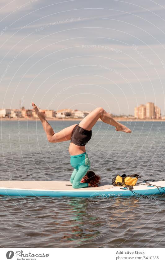 Woman doing yoga with headstand and spread legs on paddleboard woman pose balance practice flexible activity sportswear wellness salamba sirsasana vitality