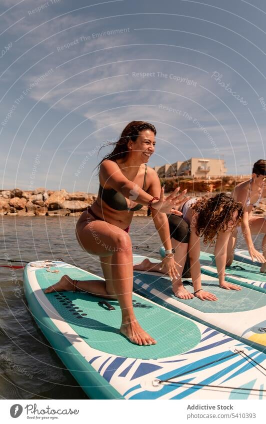 Young women doing yoga exercise on paddleboard on sea practice fitness balance wellbeing virabhadrasana ocean summer happy group arms raised training wellness
