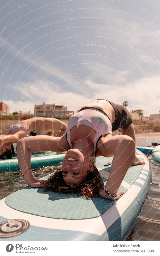 Unrecognizable women doing yoga on paddleboard in daylight girlfriend practice half wheel flexible balance swimwear seawater young female cloudy blue sky summer