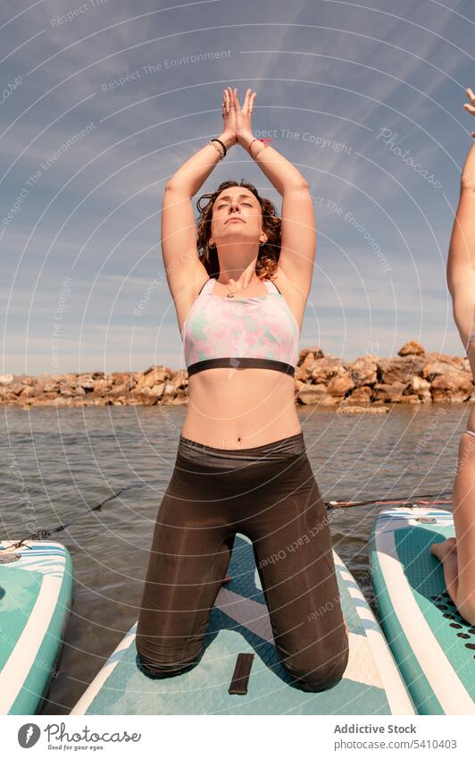 Young woman doing yoga pose on paddleboard friend eyes closed mudra stretch kneel mountain with arms up young female summer cloudy blue sky daytime vacation