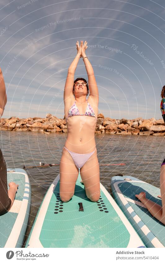 Young woman doing yoga pose on paddleboard friend eyes closed mudra stretch kneel mountain with arms up young female summer cloudy sunglasses blue sky vacation