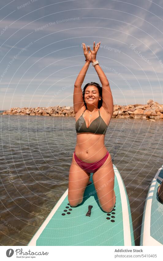 Young woman doing yoga pose on paddleboard friend eyes closed mudra stretch kneel mountain with arms up smile young female cheerful summer cloudy blue sky