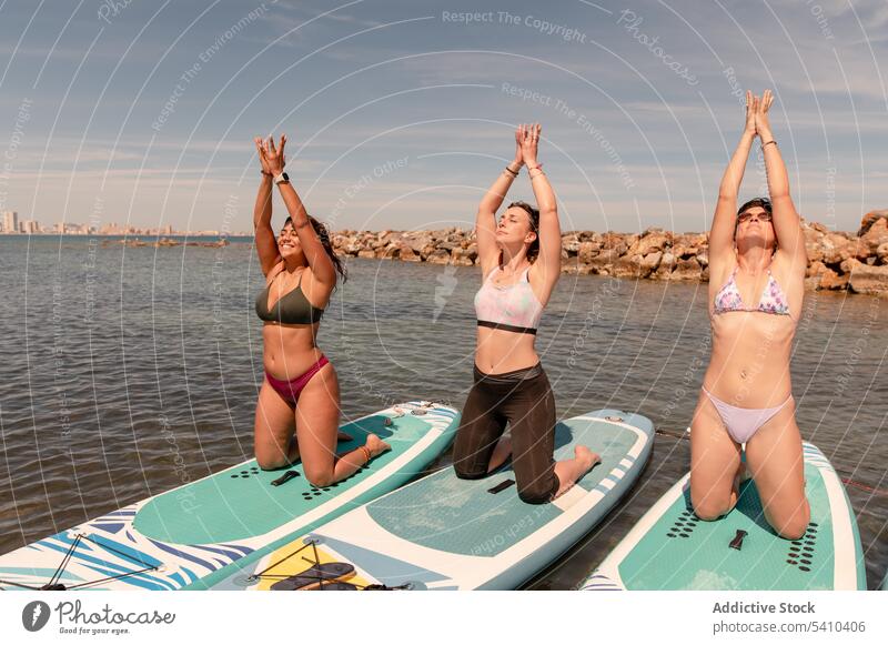 Group of young women doing yoga pose on paddleboards group mountain with arms up eyes closed mudra stretch kneel female friend summer cloudy blue sky daytime