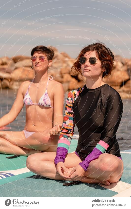 Young women sitting in Padmasana pose on paddleboards yoga lotus meditate practice calm sunglasses concentrate young female summer recreation zen vacation