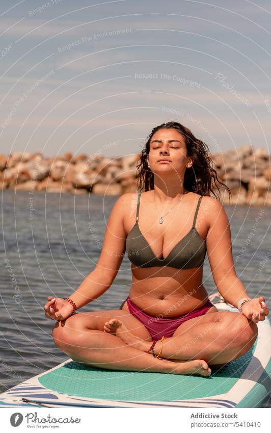 Slim woman in bikini meditating on paddleboard in sea sup yoga gyan mudra sup board meditate balance zen seaside female practice calm harmony pose wellness