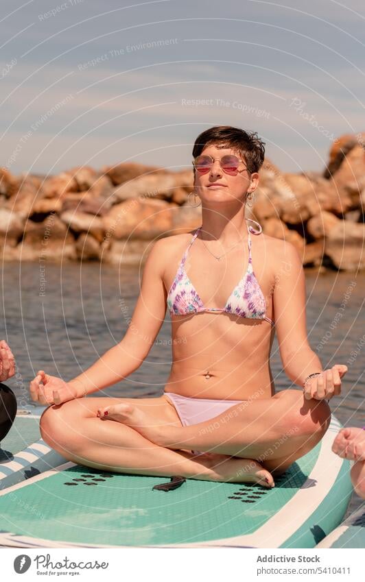 Slim woman in bikini meditating on paddleboard in sea sup yoga gyan mudra sup board meditate balance zen seaside female practice calm harmony pose sunglasses