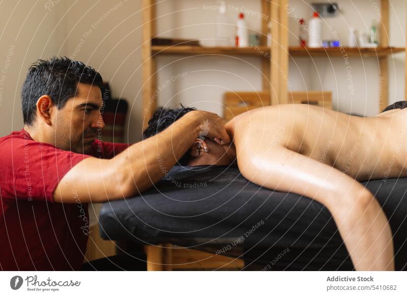 Ethnic adult masseur massaging shoulders of lying anonymous woman client in salon massage topless procedure wellness sensual therapy male hispanic ethnic sit
