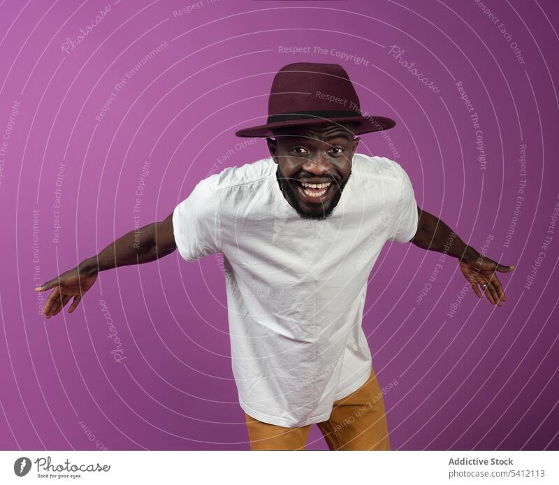Joyful black man in hat standing with outstretched arms near purple wall smile happy excited friendly freedom positive glad toothy smile male young