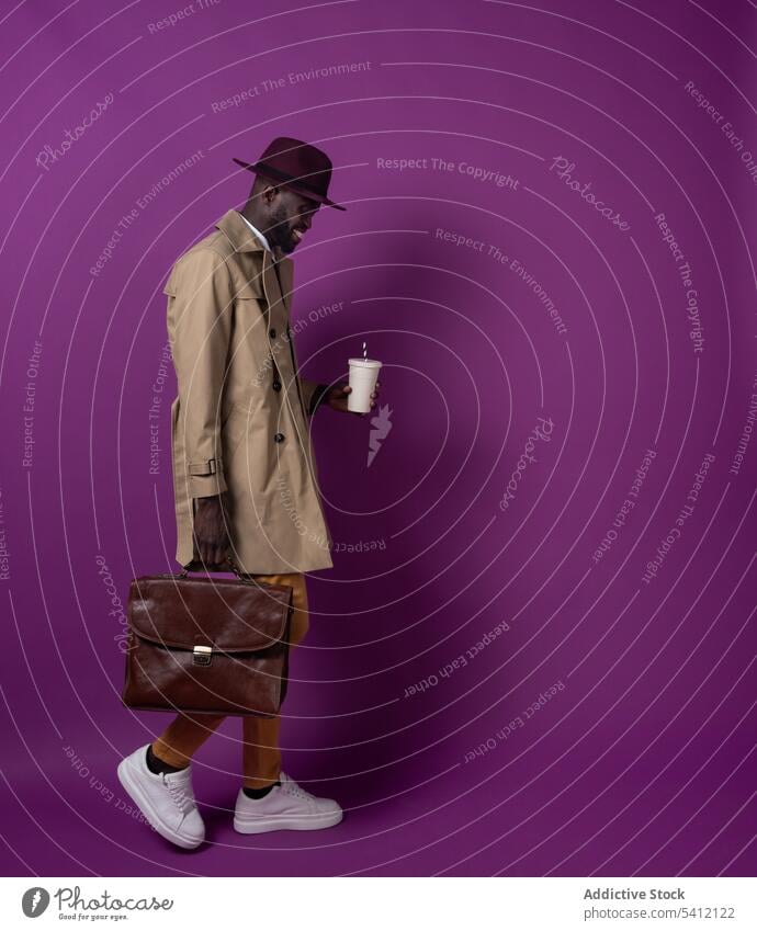 Happy young black man with briefcase and takeaway coffee cup walking near purple wall smile hot drink beard positive male african american ethnic style fashion