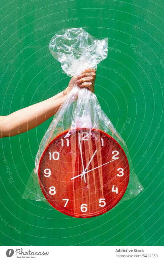 Crop person holding plastic bag with red clock in studio hand time concept show transparent wall green round package minute demonstrate material shape minimal