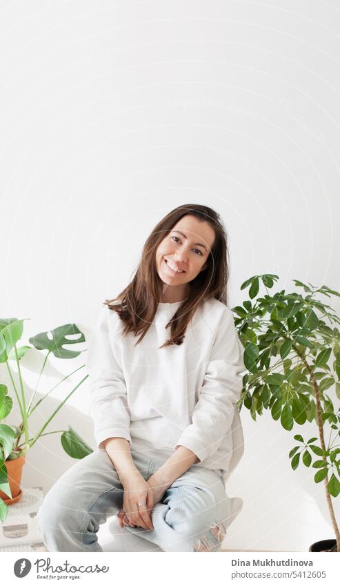 millennial woman wearing white sweatshirt and jeans, smiling, sitting on a chair at home with green plants. Hipster millennial lifestyle. Casual portrait. adult