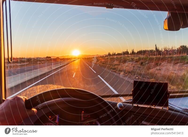 View from inside the cab of a truck driving on a highway with the early morning sun behind some mountains. interior dashboard sunset vehicle transport road