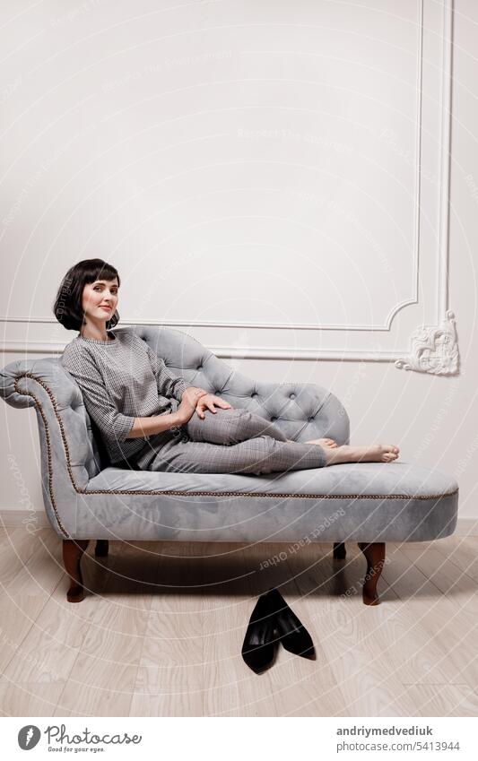 Portrait of positive young brunette female sitting barefooted on gray sofa with legs outstretched in gray checkered suit looking at camera. business woman took off her shoes and resting after work.