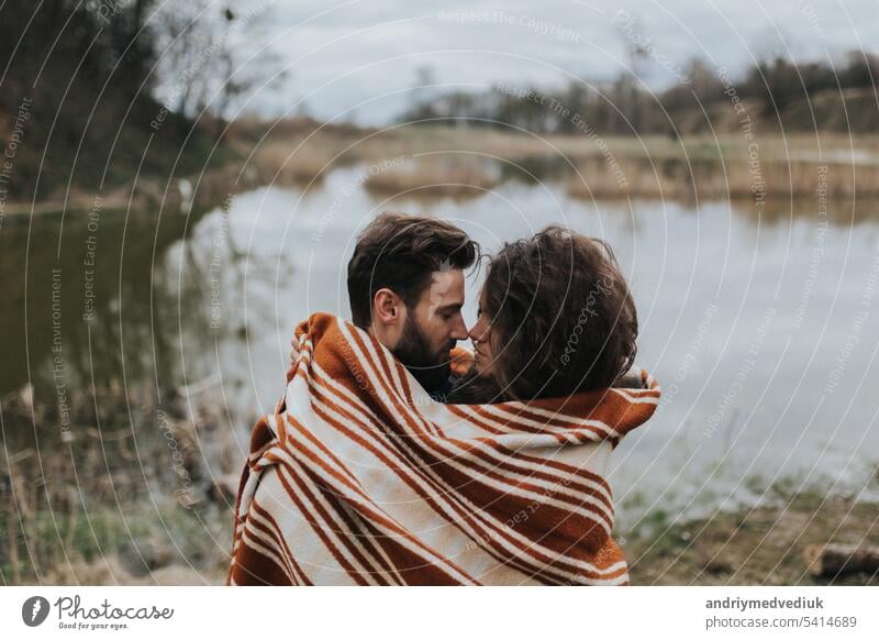 two caucasian lovers wrapped in blanket by the lake. Young couple is hugging on autumn day outdoors. A bearded man and curly woman in love. Valentine's Day. Concept of love and family.