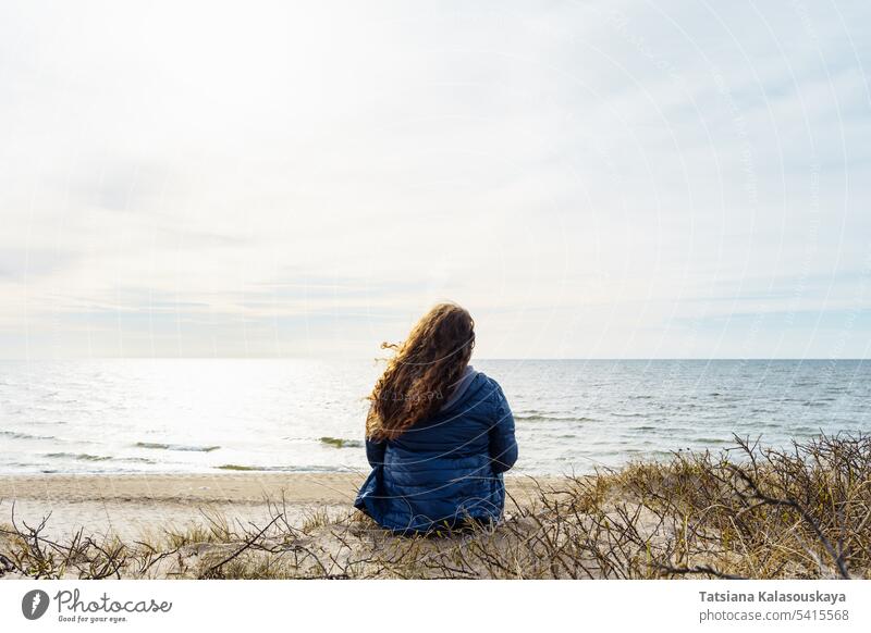 Long-haired curly plus size woman on the seashore, rear view Curly-haired thin down jacket Baltic Sea evening wind fluttering nature beauty beach coastline