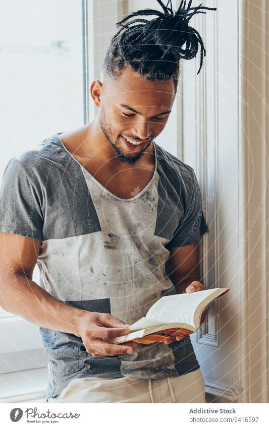 Ethnic male standing by window and reading man ethnic hairstyle braid pigtails creative book young african american person handsome attractive thoughtful