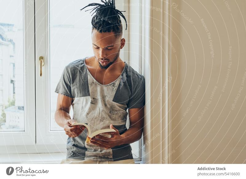 Ethnic male standing by window and reading man ethnic hairstyle braid pigtails creative book young african american person handsome attractive thoughtful