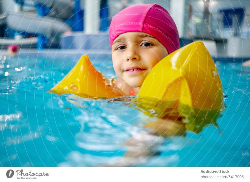 Cheerful ethnic girl in hat smiling and swimming in pool in water park leisure kid child fun two fingers peace victory joy rest relax amusement swimwear