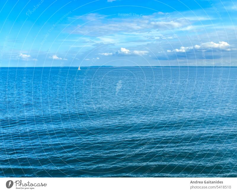 Blue image, two thirds Baltic Sea, one third slightly cloudy sky, top left a small single white sailboat, in the foreground slight waves, horizontal White Lake