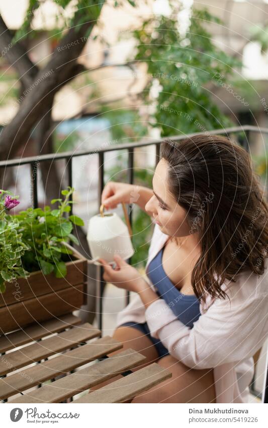 Beautiful woman watering green plants on the balcony closeup. Happy brunette smiling. Plants and flowers gardening hobby. Urban jungle millennial home interior apartment.