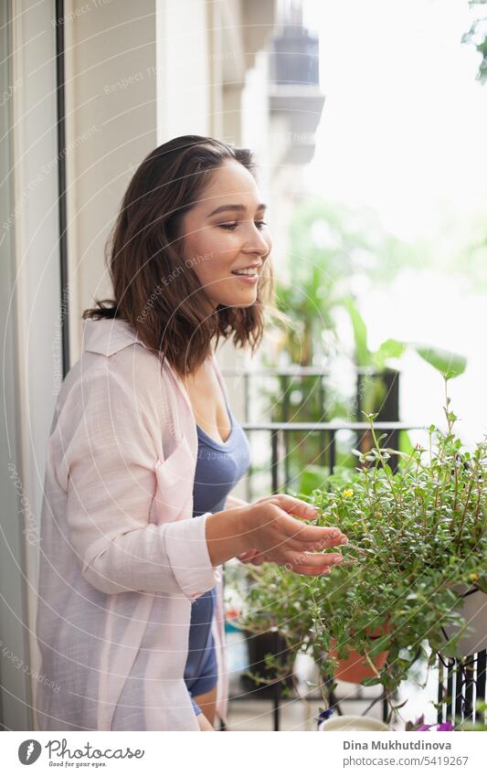 Beautiful woman smiling, taking care of green plants on the balcony closeup. Happy brunette smiling. Plants and flowers gardening hobby. Urban jungle millennial home interior apartment.