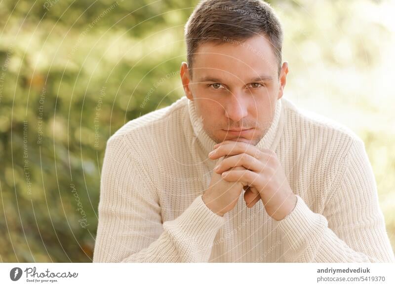 Portrait of young man in white knit casual jumper folds hands under chin looks confidently at camera on autumn nature background in countryside or in park. Concept of style